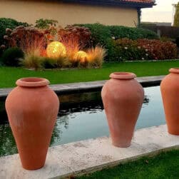 Tre høye terracottakrukker plassert på rekke langs et vannspeil i en velstelt hage med gress, busker og varm belysning i bakgrunnen.